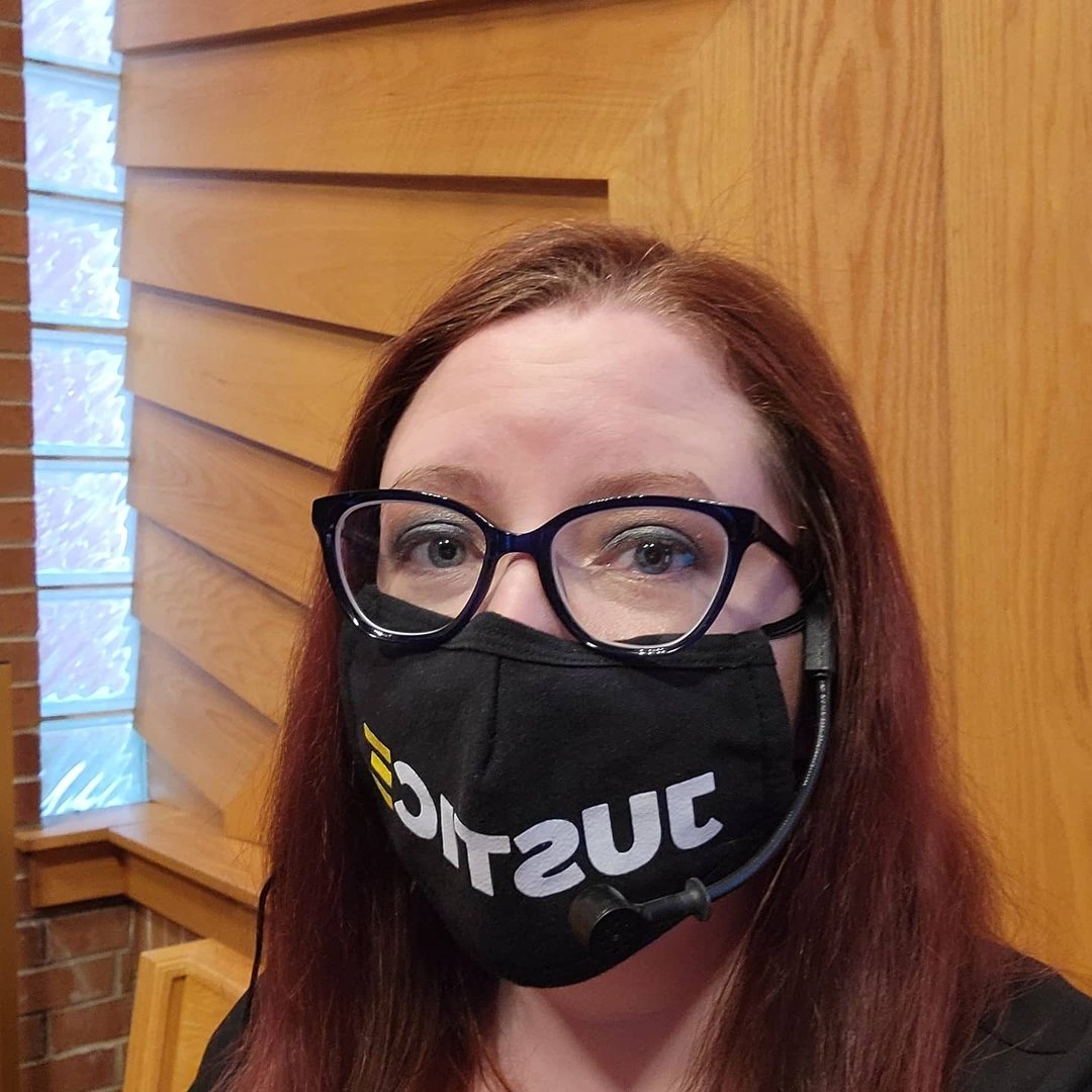 A white woman with red hair and blue glasses faces the camera, wearing a black face mask that says "Justice" across it.